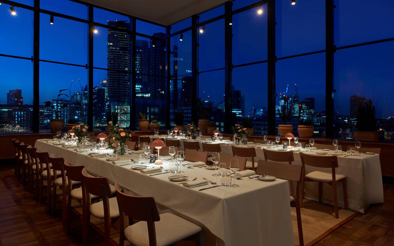 Elegant dining area with pink lamps and flowers on long tables, overlooking a city skyline through large windows at night.