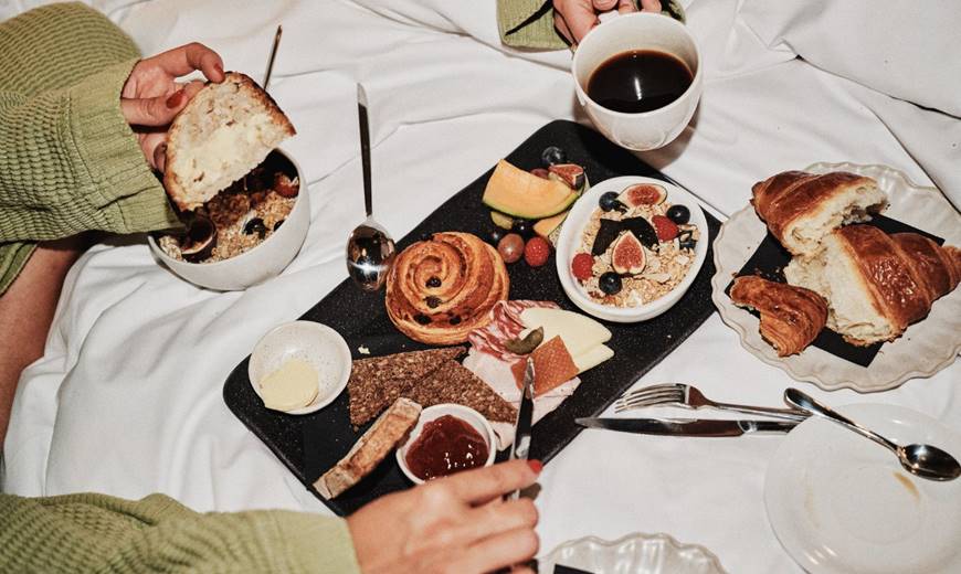 A breakfast spread on a bed, including fresh fruit, pastries, granola, cheese, bread, and coffee, with hands reaching in.