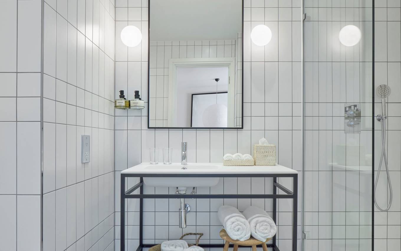 White-tiled hotel bathroom with a minimalist sink, rolled towels, and woven baskets on the black and white tiled floor.