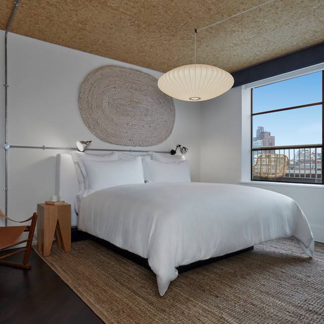 Minimalist hotel room in London with a white bed, woven wall art, and a large window showcasing a view of the city.