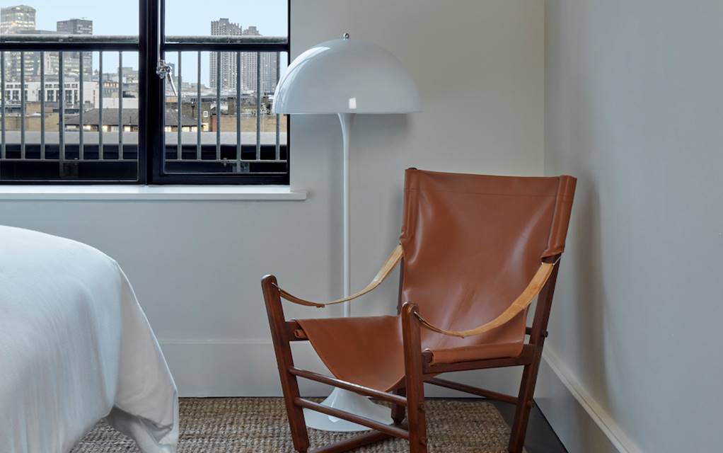 Contemporary hotel room in London with leather lounge chair, white floor lamp, and window view to the city.