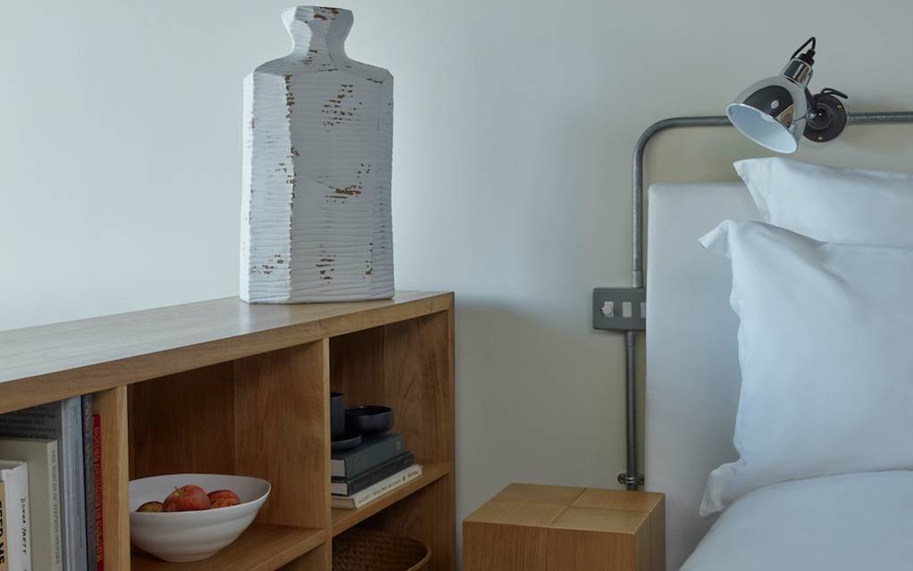 Minimalist bedside with white bedding, a metal reading light, and a wooden bookshelf with a vase in a hotel room in London.