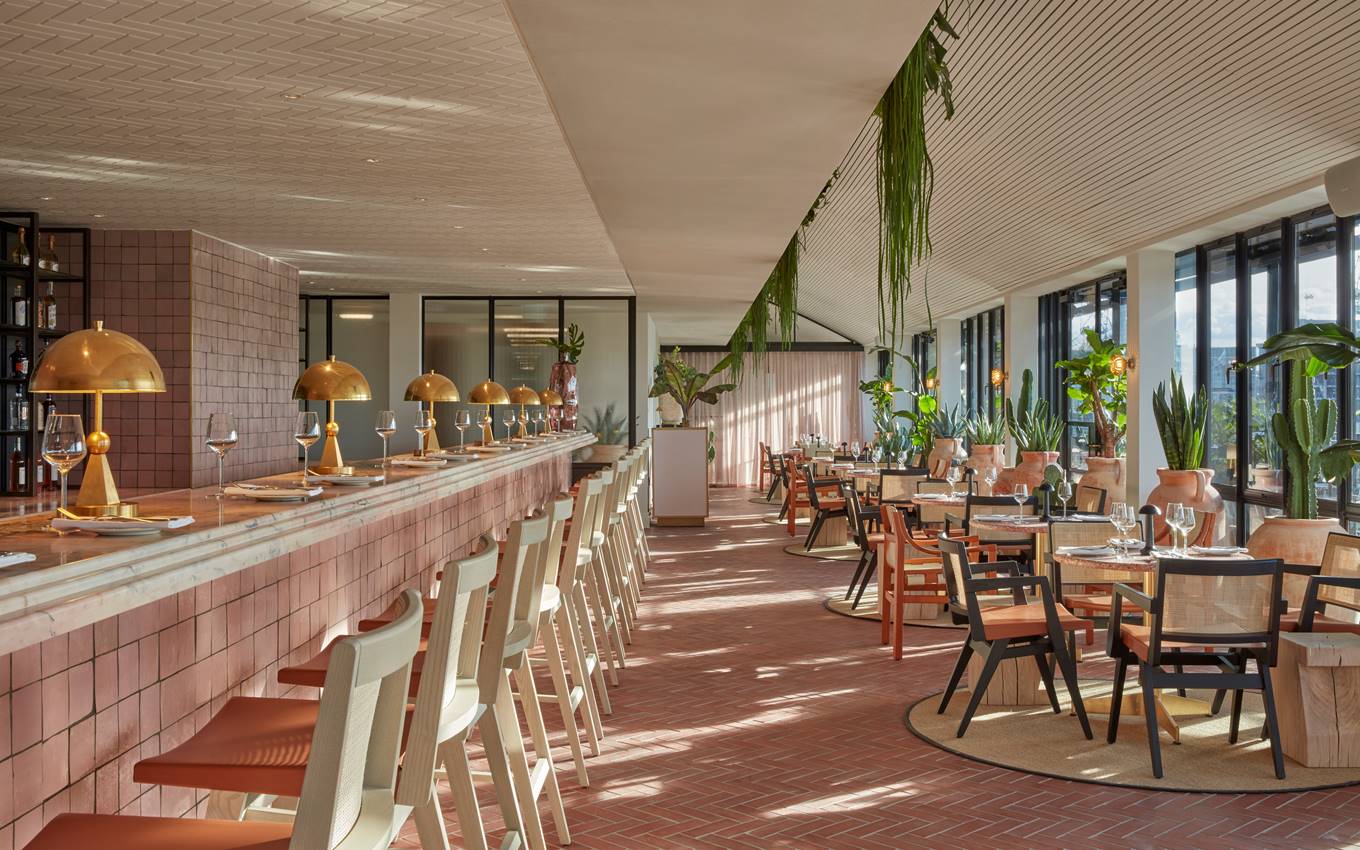 Spacious London restaurant with a long bar, brass lamps, and tables set for dining, decorated with plants and natural light.