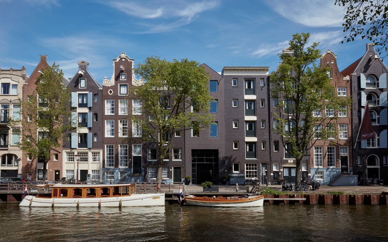 Exterior of the Pulitzer Amsterdam hotel with tall windows, trees in the front and two boats docked by the water.