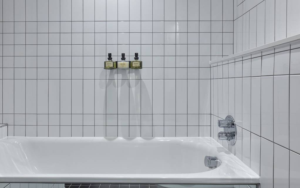 Modern white tiled bathtub with mounted bath products against the wall in a hotel room at One Hundred Shoreditch in London.