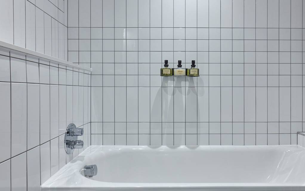 Modern white tiled bathtub with mounted bath products against the wall in a hotel room at One Hundred Shoreditch in London.