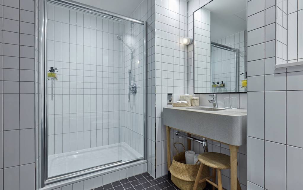Tiled hotel bathroom at One Hundred Shoreditch with glass shower, vanity, and storage baskets.