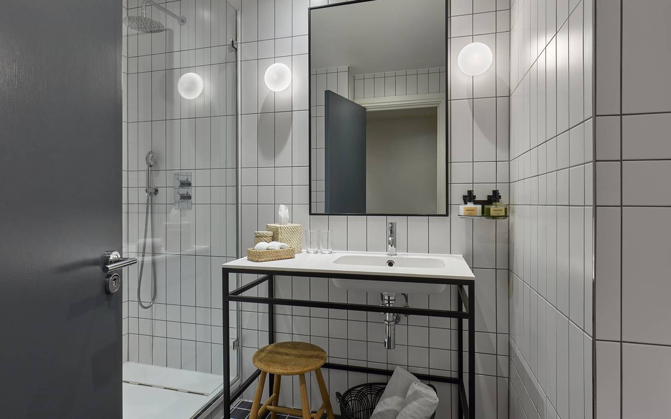 Minimalist bathroom at a London hotel with white tiles, modern sink on a black frame, and a small wooden stool.