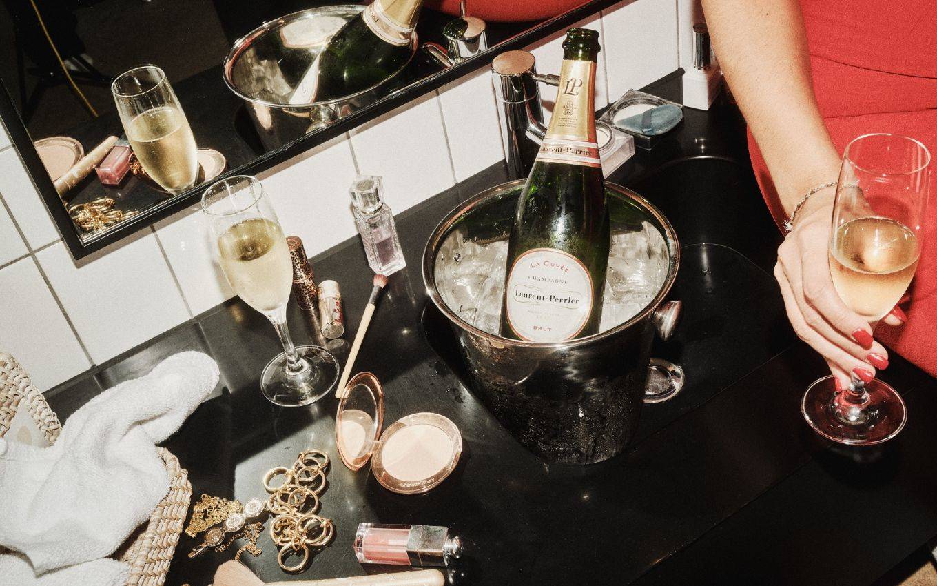 A bottle of Laurent-Perrier in an ice bucket sits on a vanity surrounded by makeup and accessories.