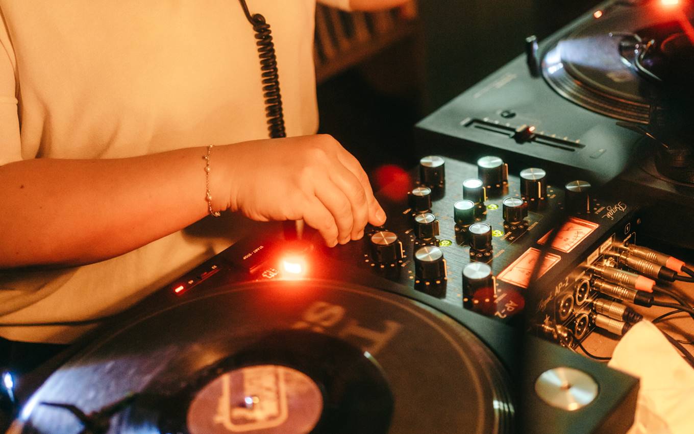 A close-up of a DJ’s hands adjusting knobs on a turntable, illuminated by warm light, with vinyl records and cables in view.