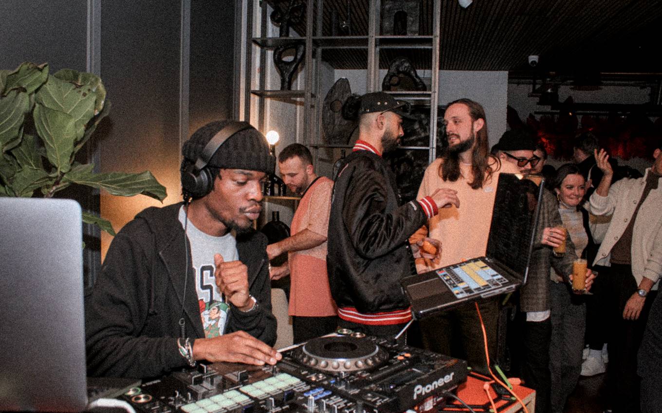 A DJ in a black beanie and headphones playing a set at One Hundred Shoreditch, while guests chat and dance nearby.