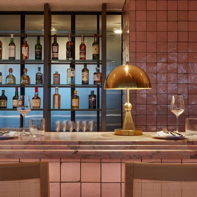 A stylish bar in London with pink tiles, a marble counter, a gold lamp, and a backlit shelf displaying a selection of spirits.