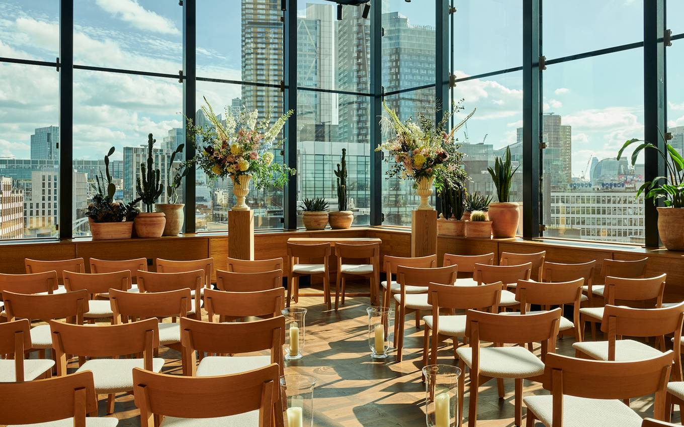 Sunlit room set for an event in London at One Hundred Shoreditch with wooden chairs, flowers, and city views through windows.