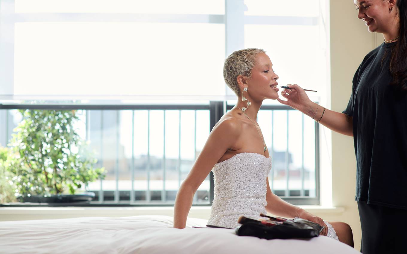 A woman in a strapless white dress receives makeup touch-ups, seated near a bright window with city views in the background.