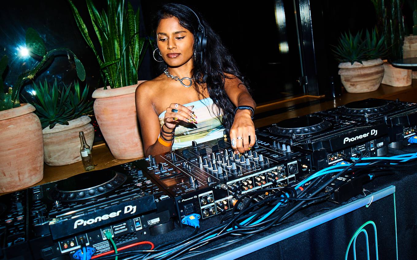 Female DJ in a strapless top and silver jewellery mixing tracks at a Pioneer DJ booth with potted plants at a London bar.