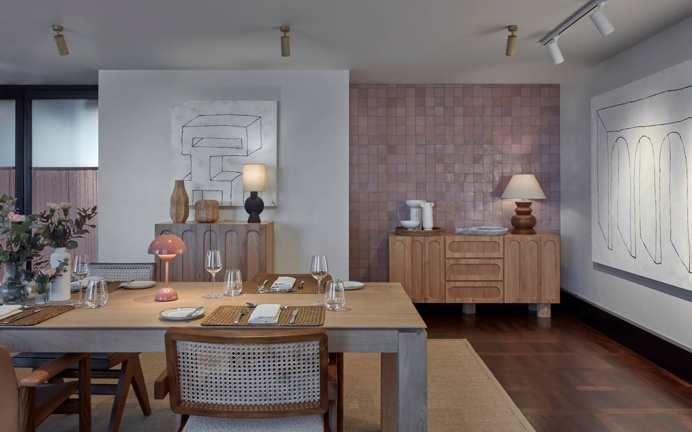 Dining area with wooden table, pink wall tiles, artistic decor, and place settings for guests, creating a cosy ambiance.