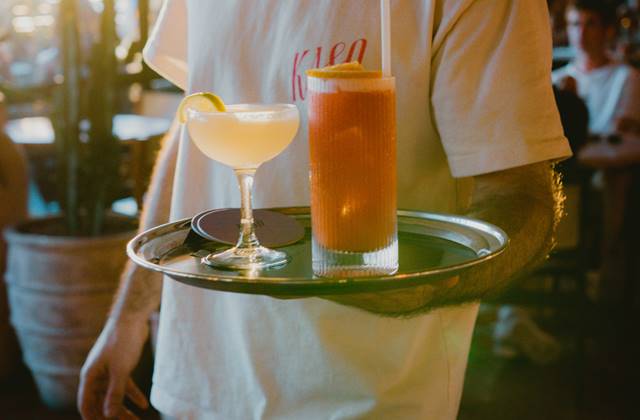 A man stands confidently, holding a tray with two drinks, ready to serve in a social setting in Kaso.