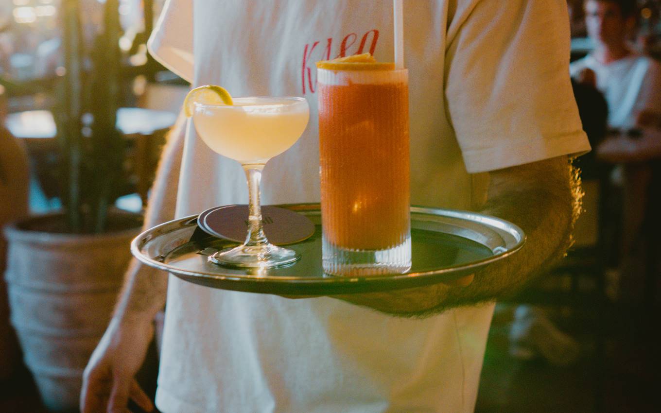 Bartender in a white t-shirt carries a tray with two colourful cocktails in a lively bar setting at Kaso.