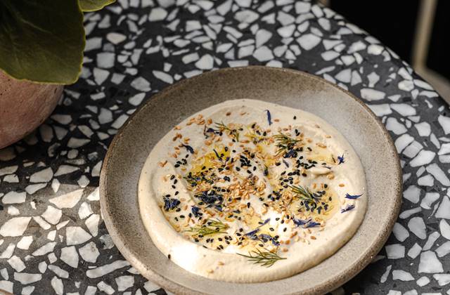 A bowl of creamy hummus sits on a table beside a vibrant green plant, creating a fresh and inviting atmosphere.