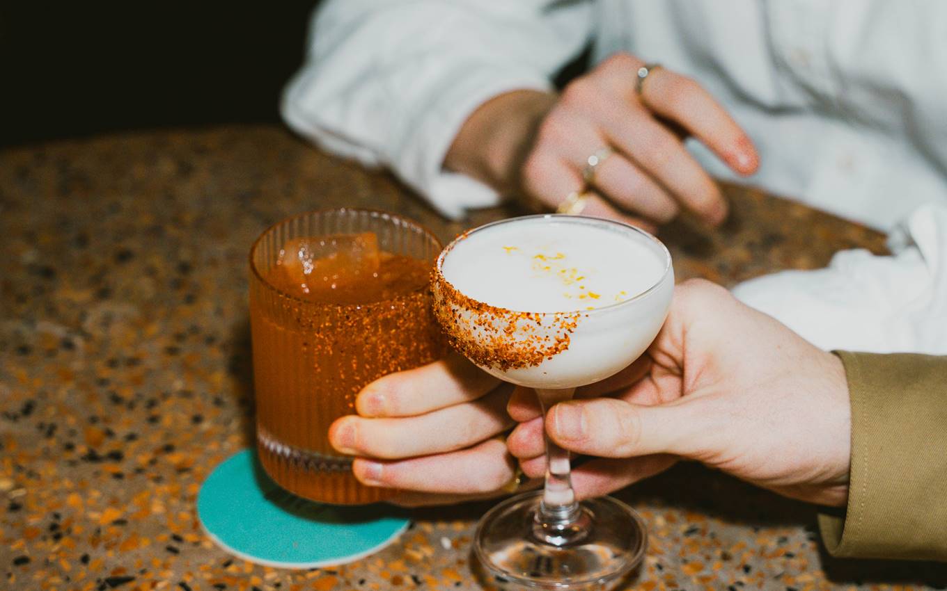Two hands holding cocktails, one with a salted rim and frothy top, the other a spicy margarita with a chilli-rimmed glass.