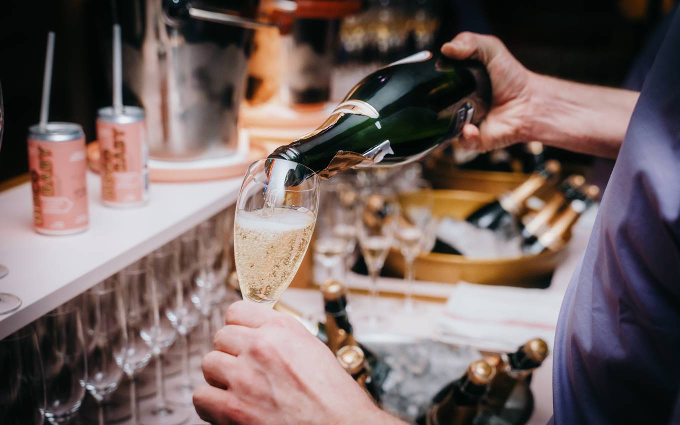 Person pouring sparkling wine into a glass at a bar setup with rows of glasses, cans, and ice buckets.