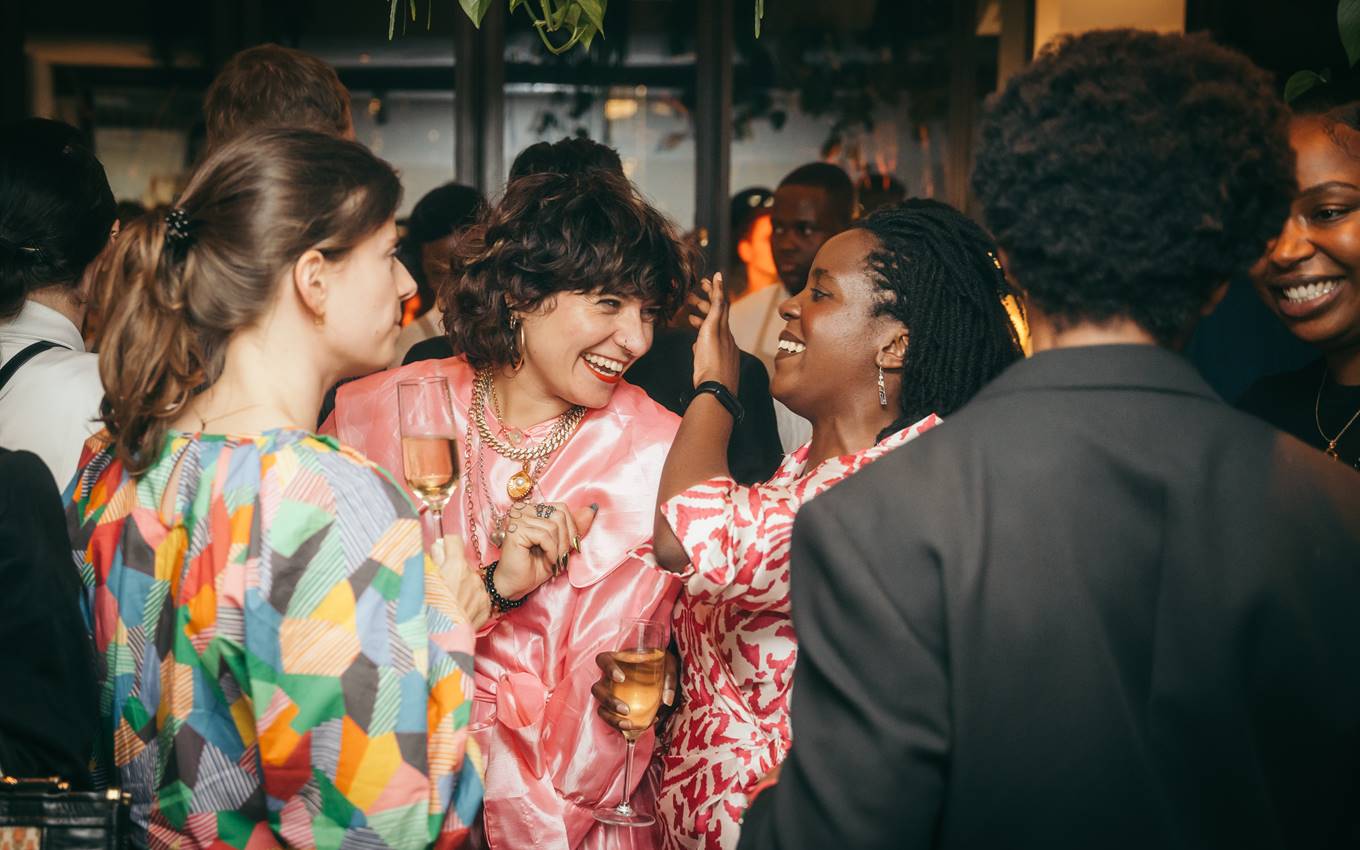 Friends at a central London party, chatting and laughing while holding champagne glasses, dressed in vibrant clothing.