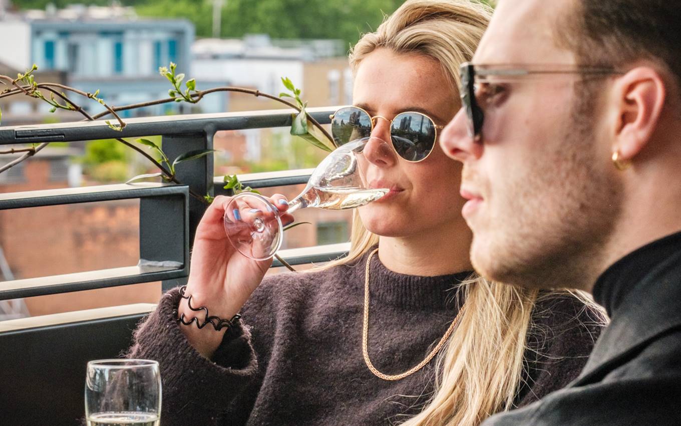 A woman sipping champagne outdoors with a man beside her, enjoying a drink on a London hotel’s rooftop terrace.