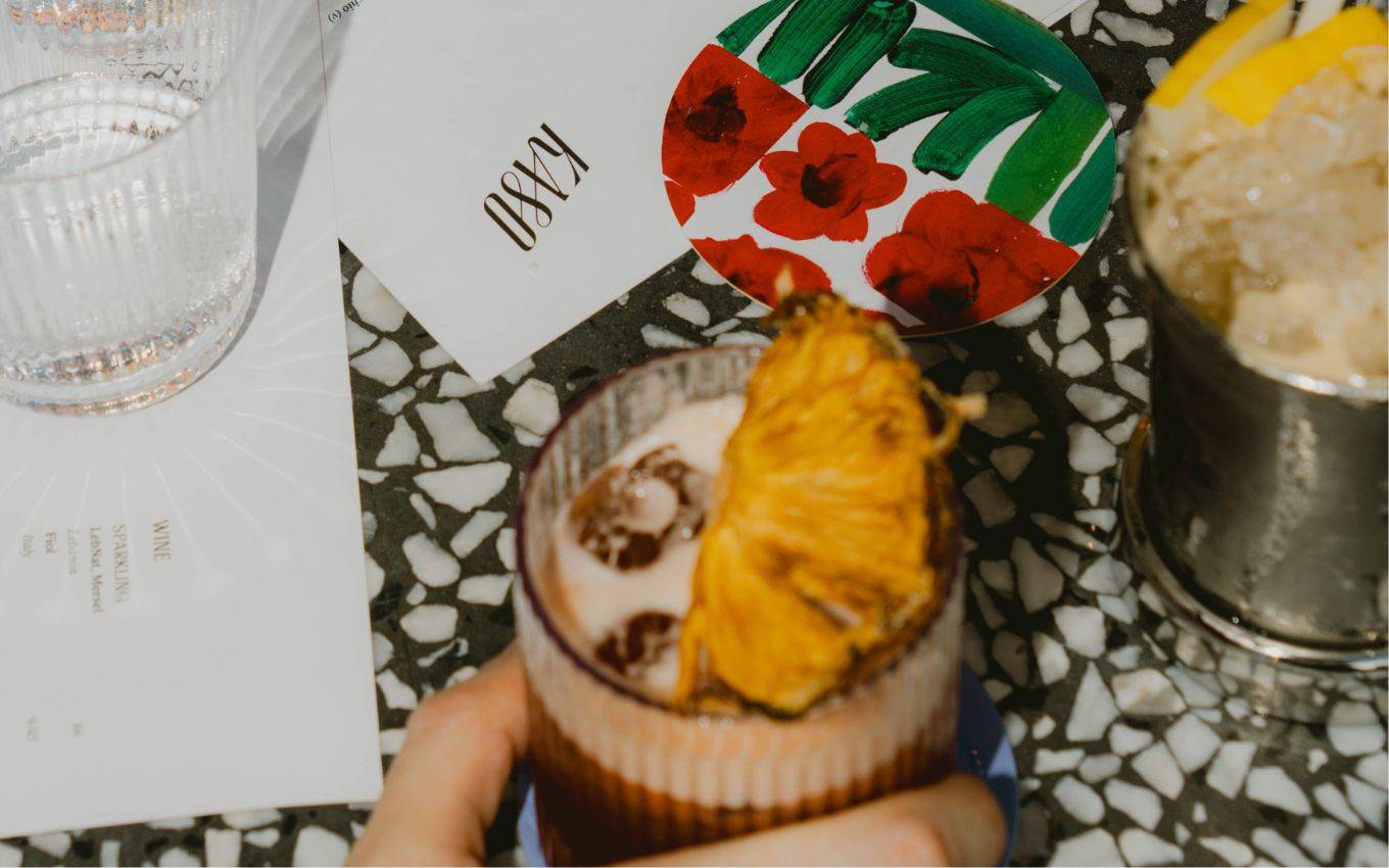 A cocktail garnished with dried pineapple is held over a terrazzo table with a floral-patterned plate and a KA80-branded napkin.