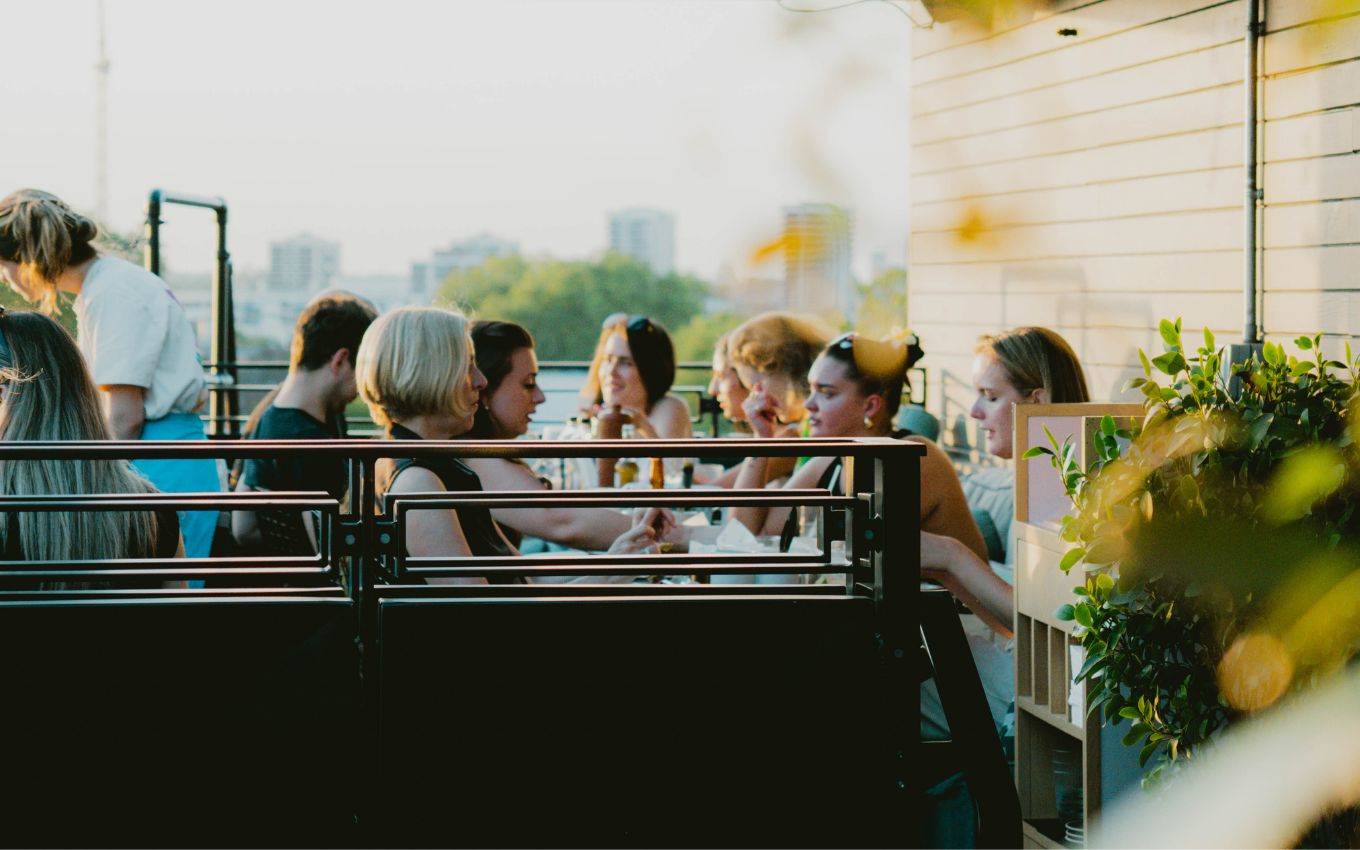 A group of people enjoying drinks and conversation on a rooftop terrace at sunset, with city buildings in the background.