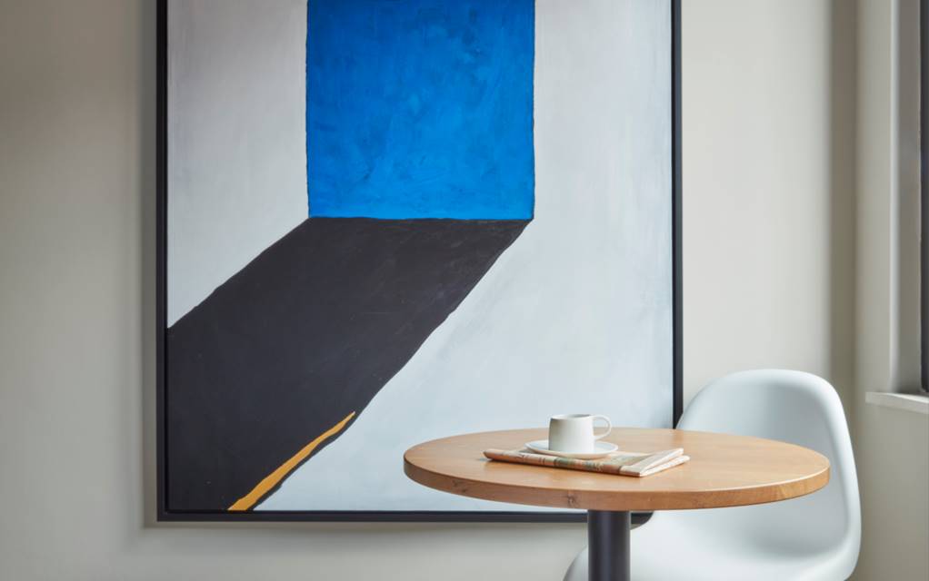 Modern art piece with bold blue and black shapes, hung beside a small wooden table and a white chair at a London hotel room.