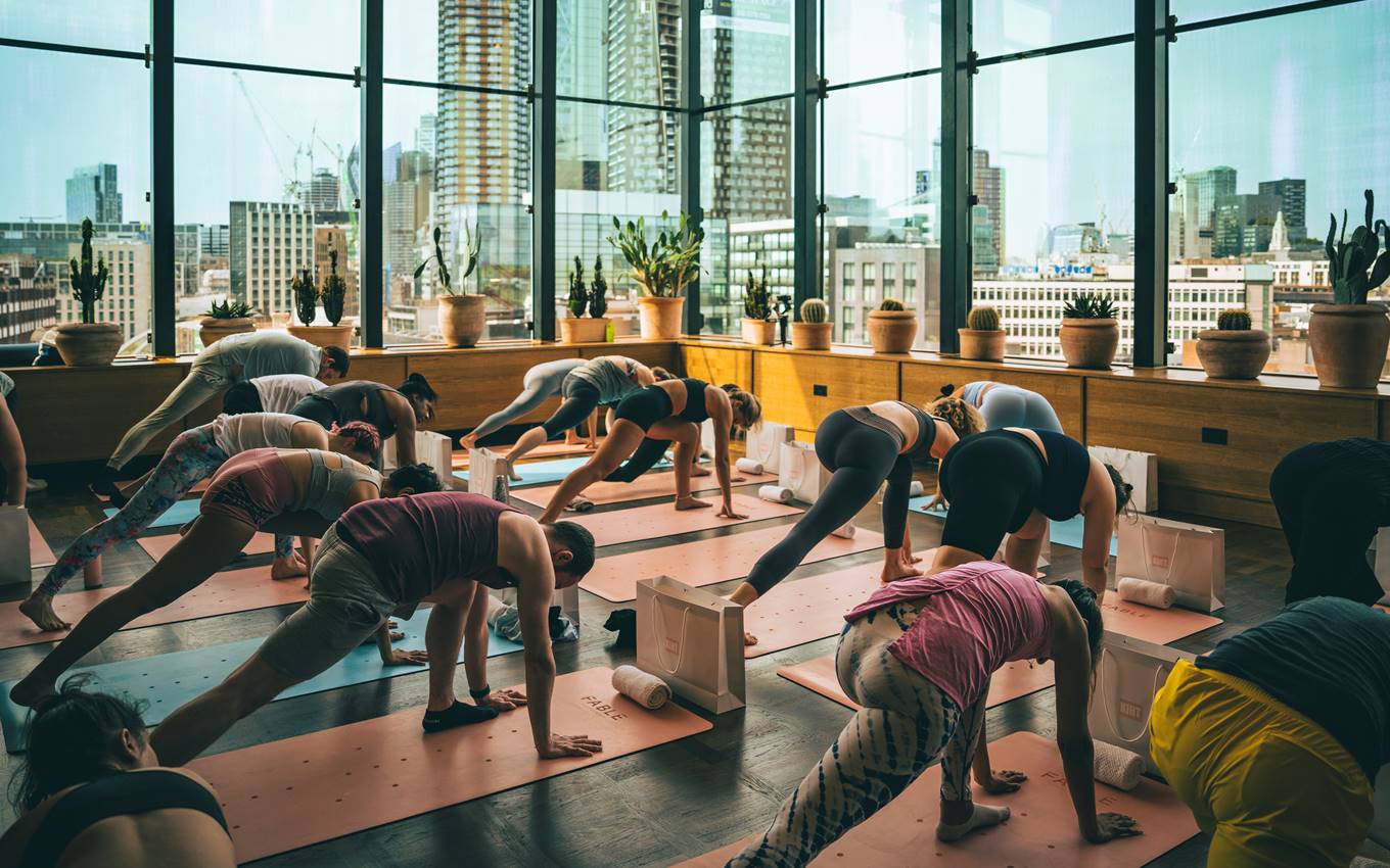 Group yoga session in a glass-walled studio filled with natural light and London city views, potted plants in the background.