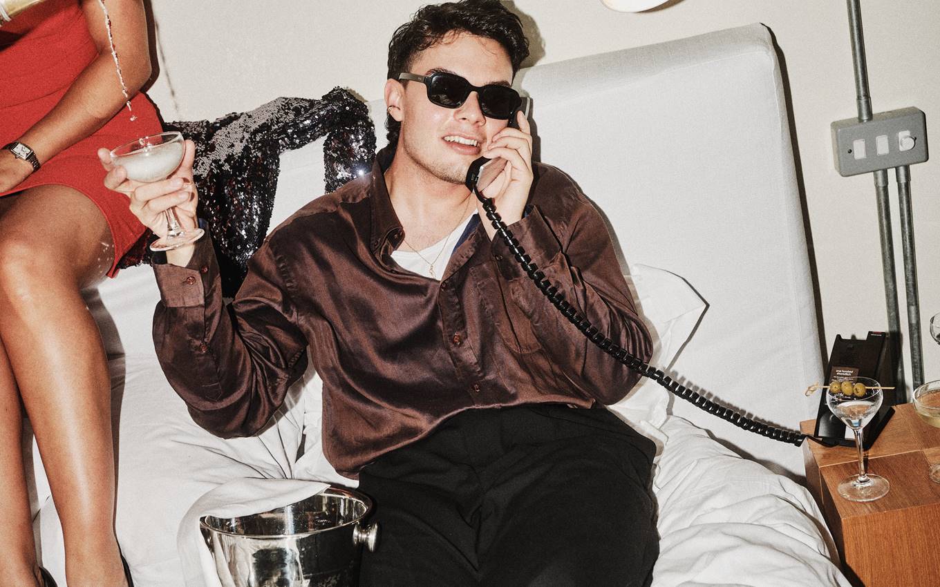 A man in sunglasses and a silk shirt lounging on a hotel bed, talking on a vintage phone and holding a cocktail.