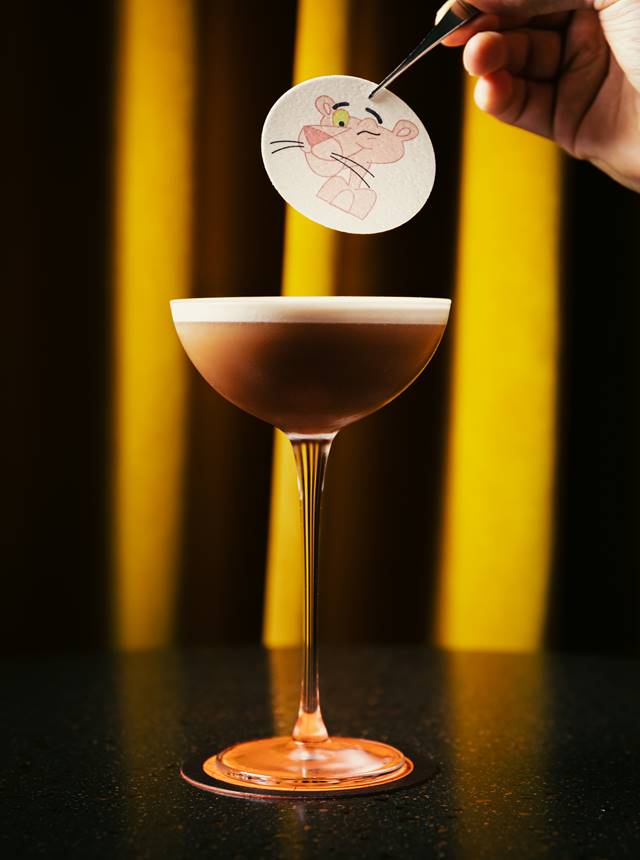 Elegant cocktail in a glass with a cartoon Pink Panther topper being placed, set against a background of yellow curtains.