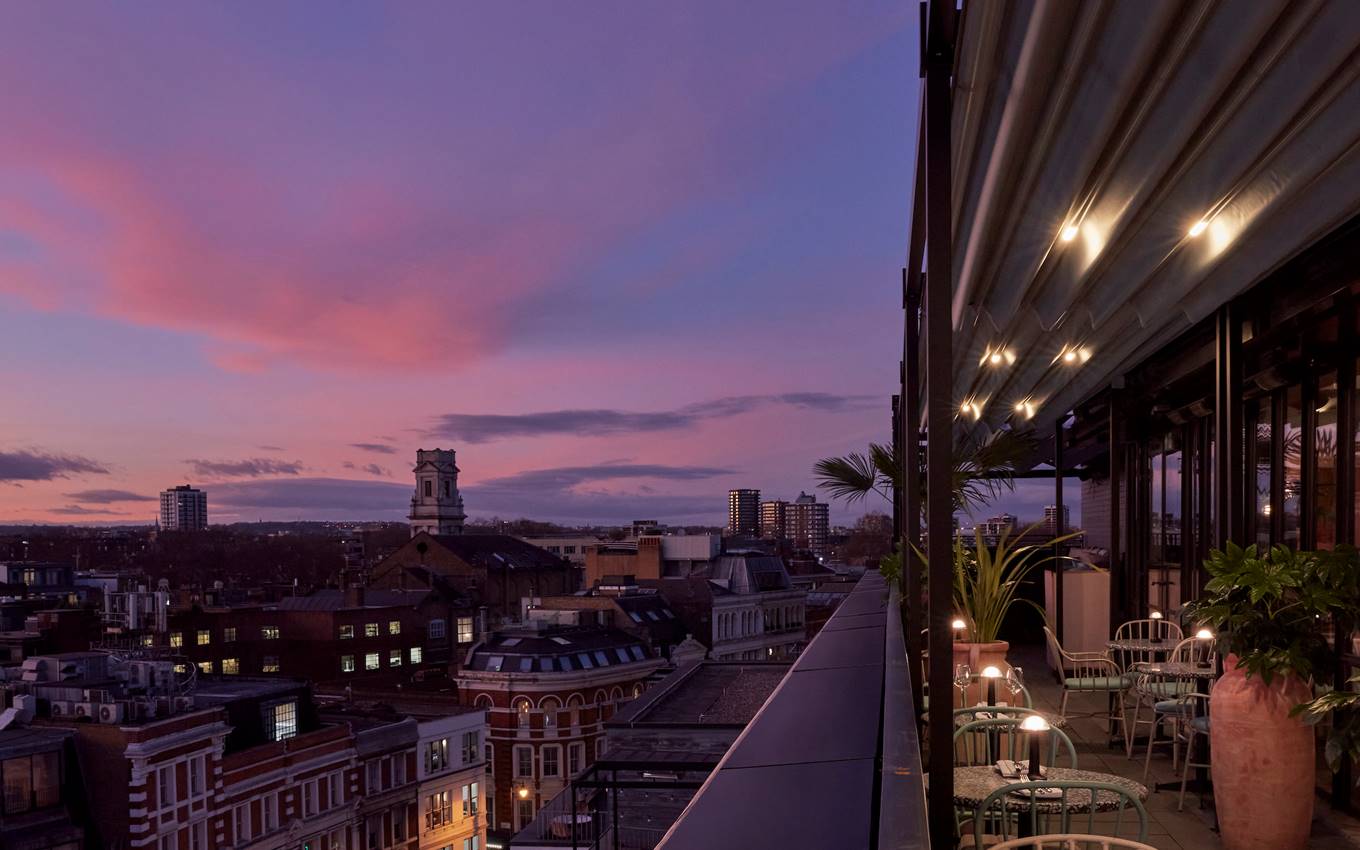 A rooftop terrace at One Hundred Shoreditch with twinkling lights, lush greenery, and a panoramic view of London at sunset.