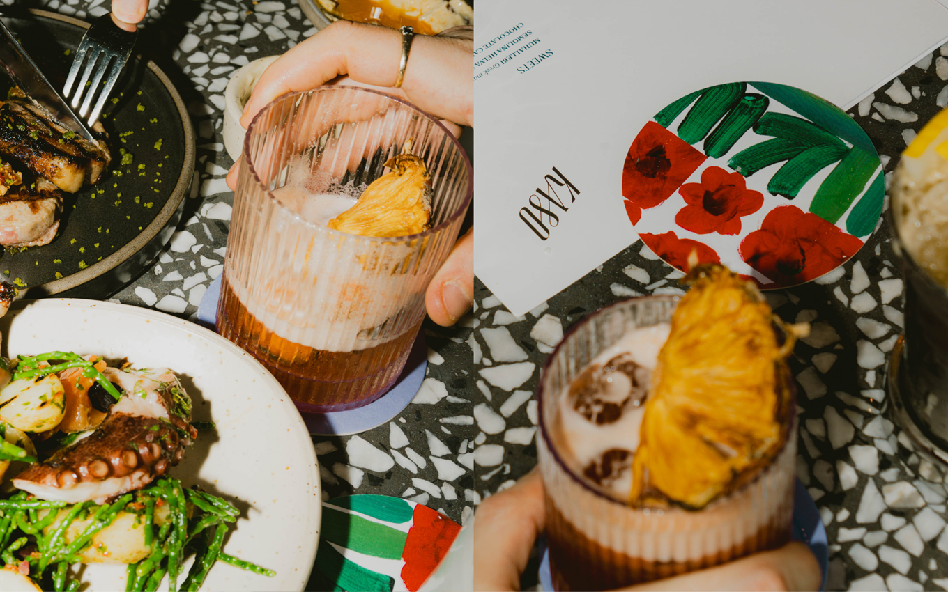 A lively dinner scene with close-up of a cocktail garnished with dried pineapple with colourful coasters and a steak dish.
